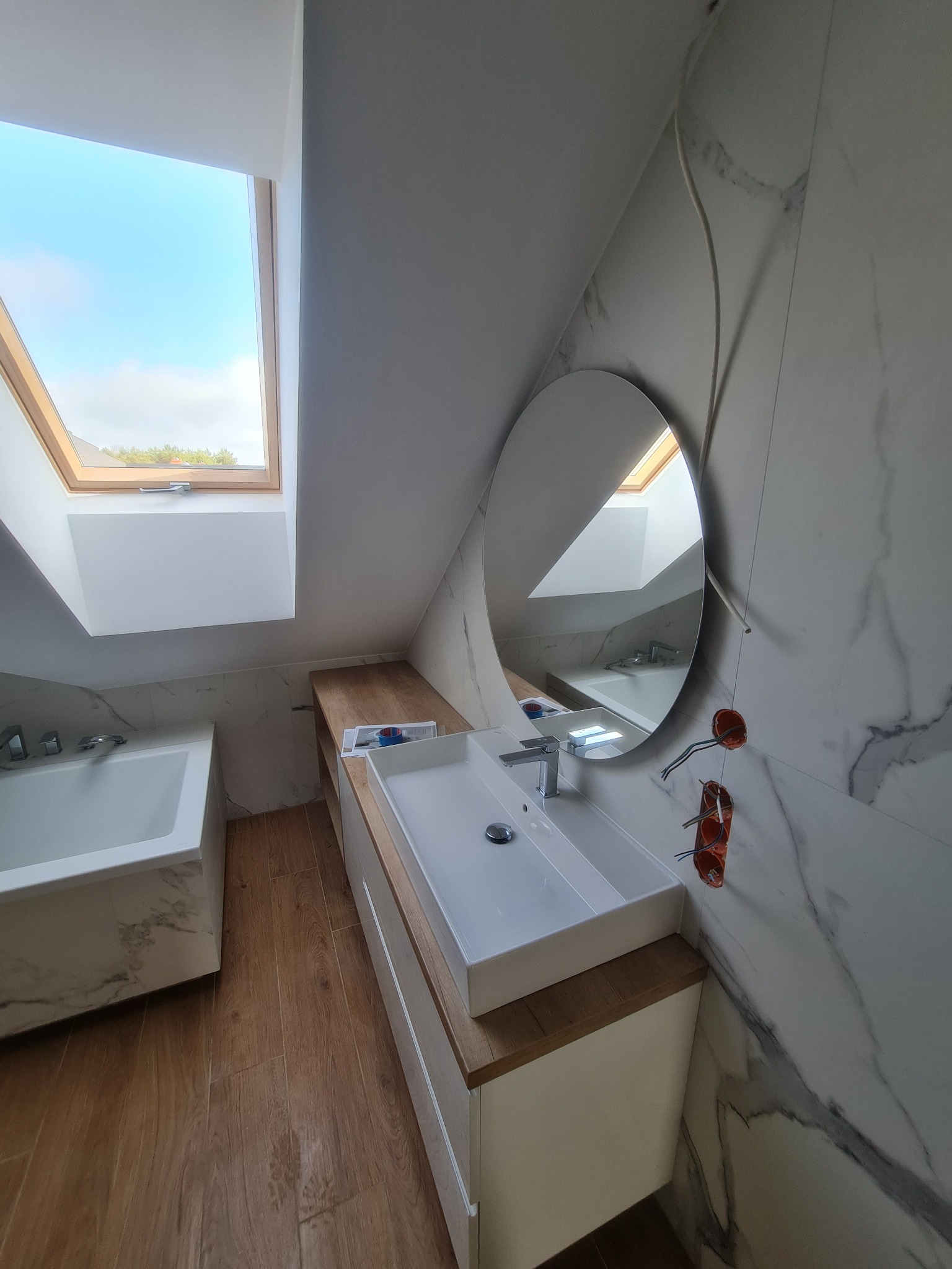 Nowoczesna łazienka na poddaszu z drewnianą podłogą, umywalką nablatową i owalnym lustrem, widoczne niezabezpieczone gniazdka elektryczne.