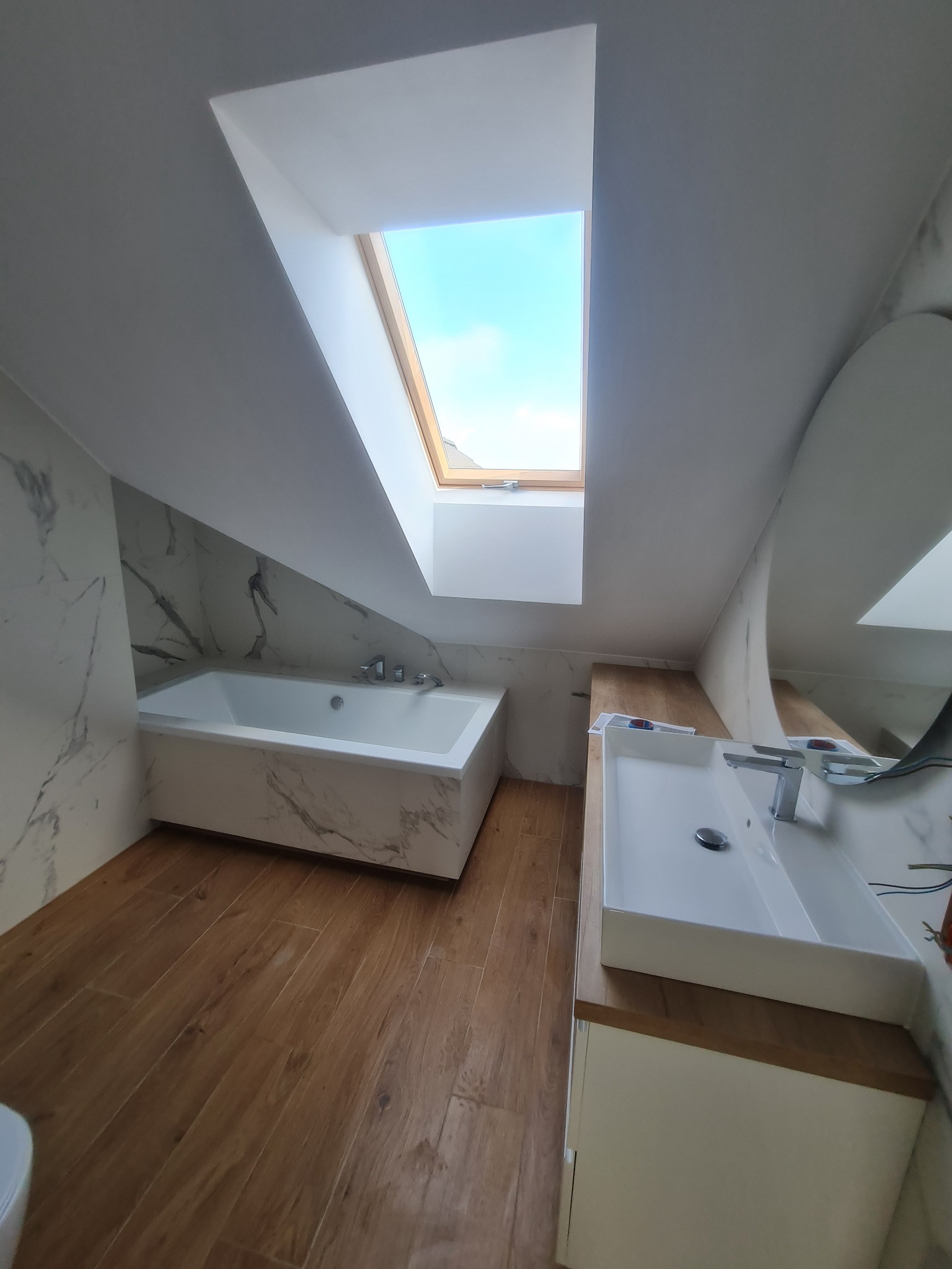 Nowoczesna łazienka na poddaszu z drewnianą podłogą, marmurową wanną i umywalką nablatową pod oknem dachowym.
