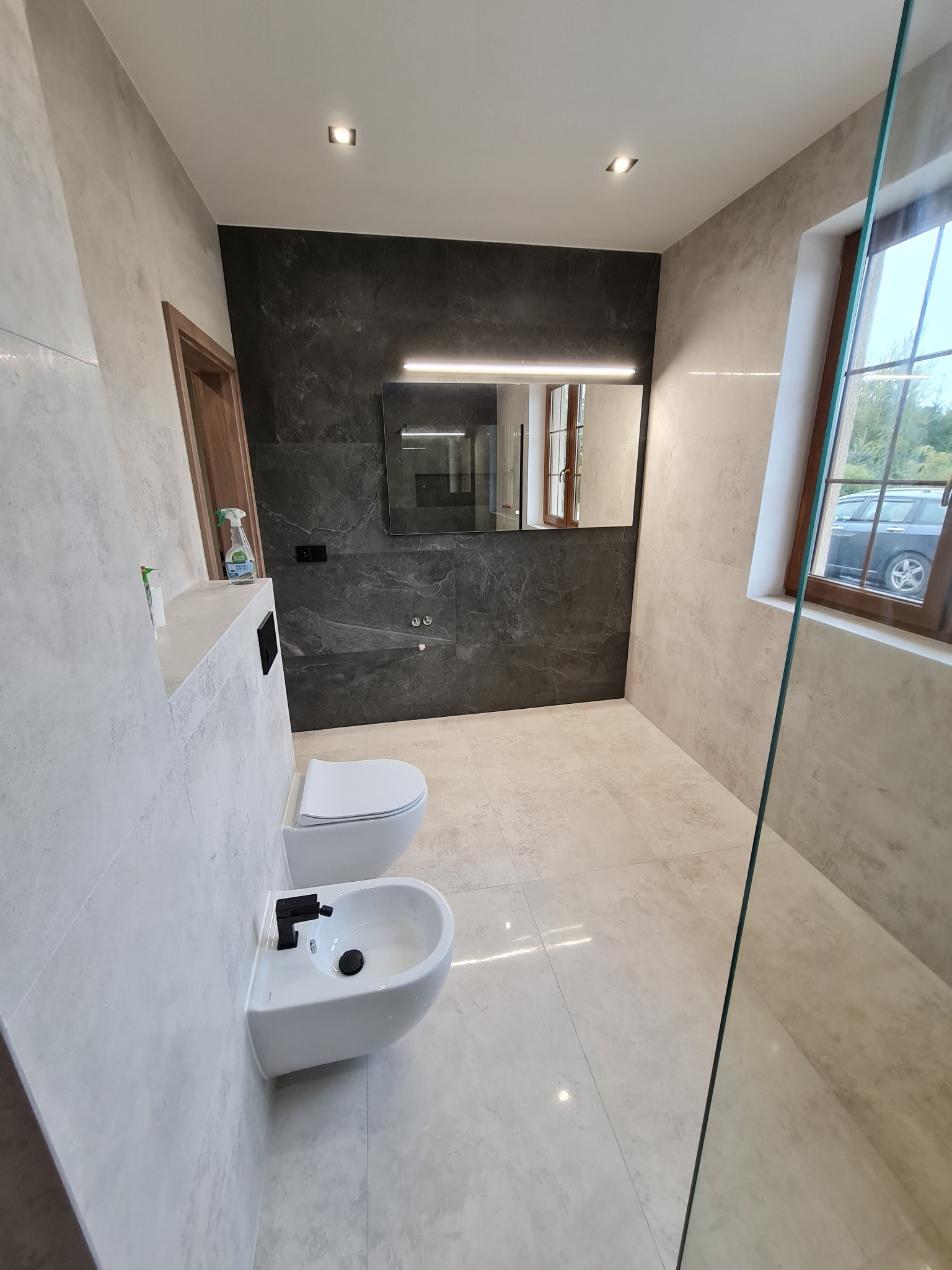 Nowoczesna łazienka z szarymi płytkami imitującymi beton, wiszącą toaletą i bidetem z czarną armaturą, lustrem z podświetleniem LED i widokiem na zieleń za oknem.