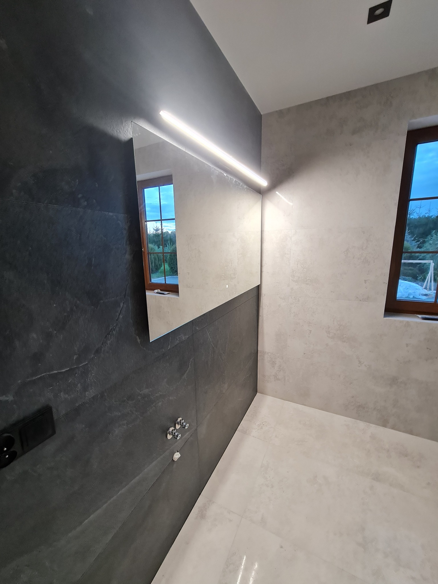 Nowoczesna łazienka z szarymi płytkami imitującymi kamień na ścianach, podświetlanym lustrem i widokiem na zewnątrz przez okno