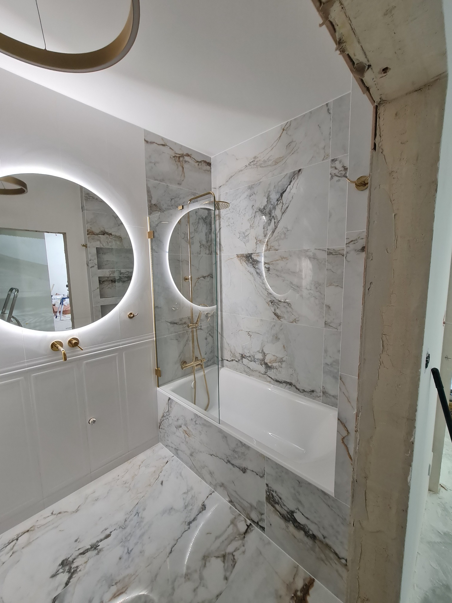 Nowoczesna łazienka w trakcie wykończenia, z białymi płytkami imitującymi marmur, okrągłym lustrem z podświetleniem LED i złotą armaturą, widoczna surowa ściana przed wykończeniem.