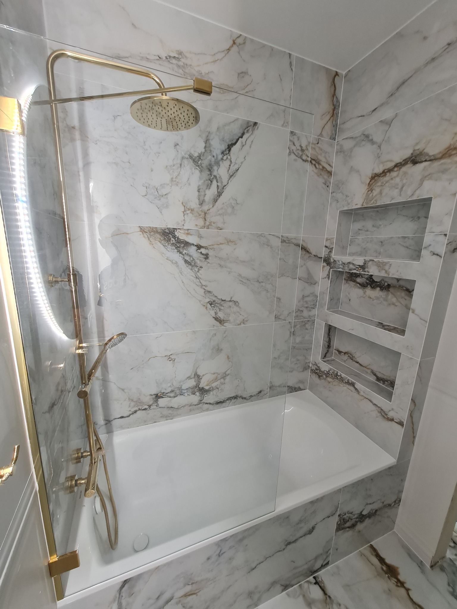 Wykończona łazienka z wanną obłożoną płytkami imitującymi marmur, z zabudowaną szklaną ścianką prysznicową z elementami w kolorze złotym oraz wnękami w ścianie.