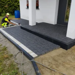 Pracownik w odblaskowej kamizelce układa ciemnoszarą kostkę brukową z jasnoszarą obwódką na podjeździe domu. Widoczny młotek brukarski, poziomica i częściowo ułożony fragment z czarnymi obrzeżami.
