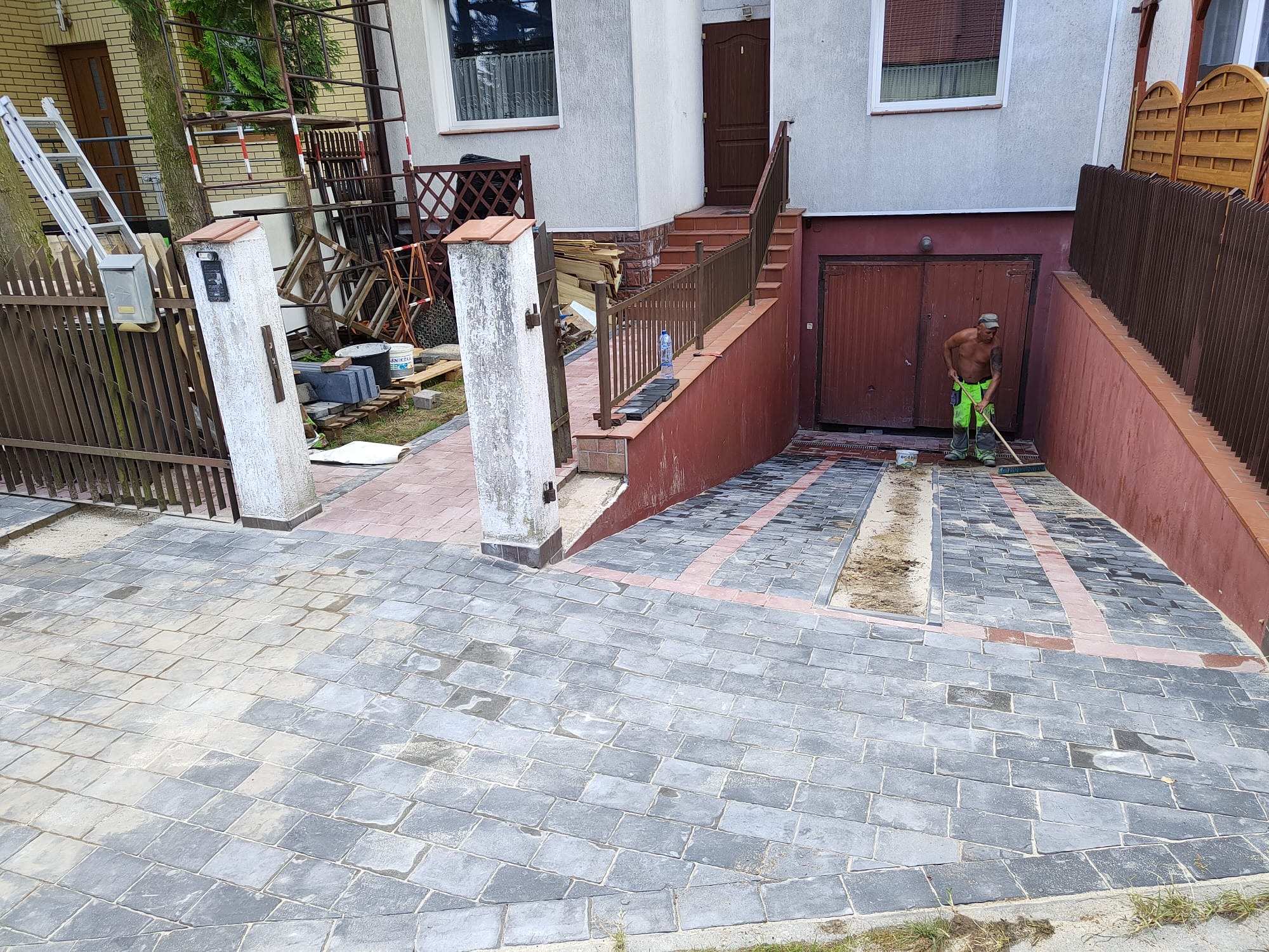 Mężczyzna w neonowo-zielonych spodniach zamiata nowo ułożoną, szarą kostkę brukową na podjeździe przed domem z brązową bramą garażową.