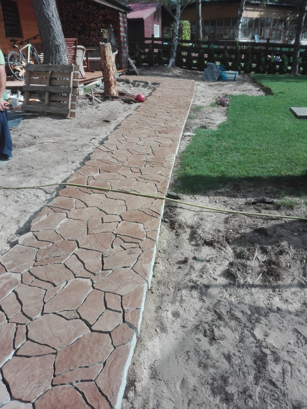 Ścieżka z nieregularnych, brązowych płyt imitujących kamień, prowadząca przez piaszczysty teren w kierunku drewnianych budynków, z widocznym fragmentem węża ogrodowego i trawnika.