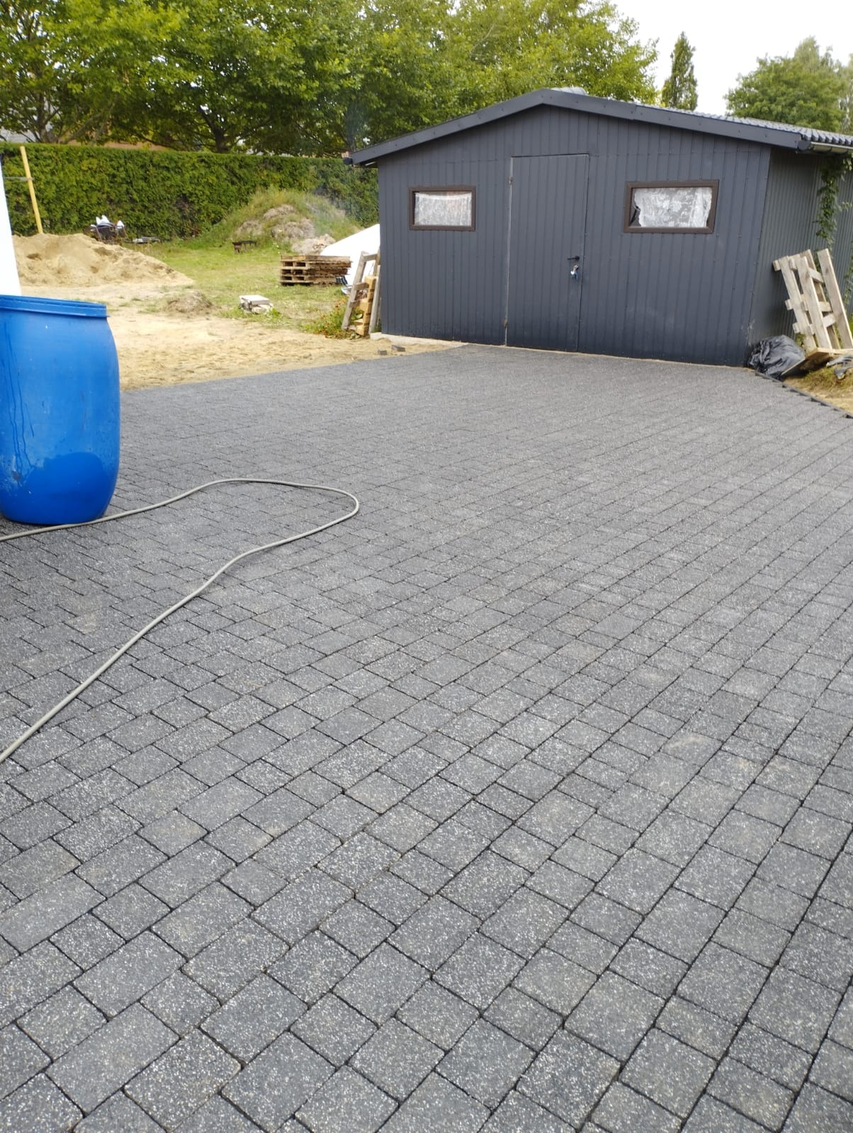 Ułożona szara kostka brukowa na podjeździe, z niebieską beczką i szarą szopą w tle, widoczne ślady prac wykończeniowych.