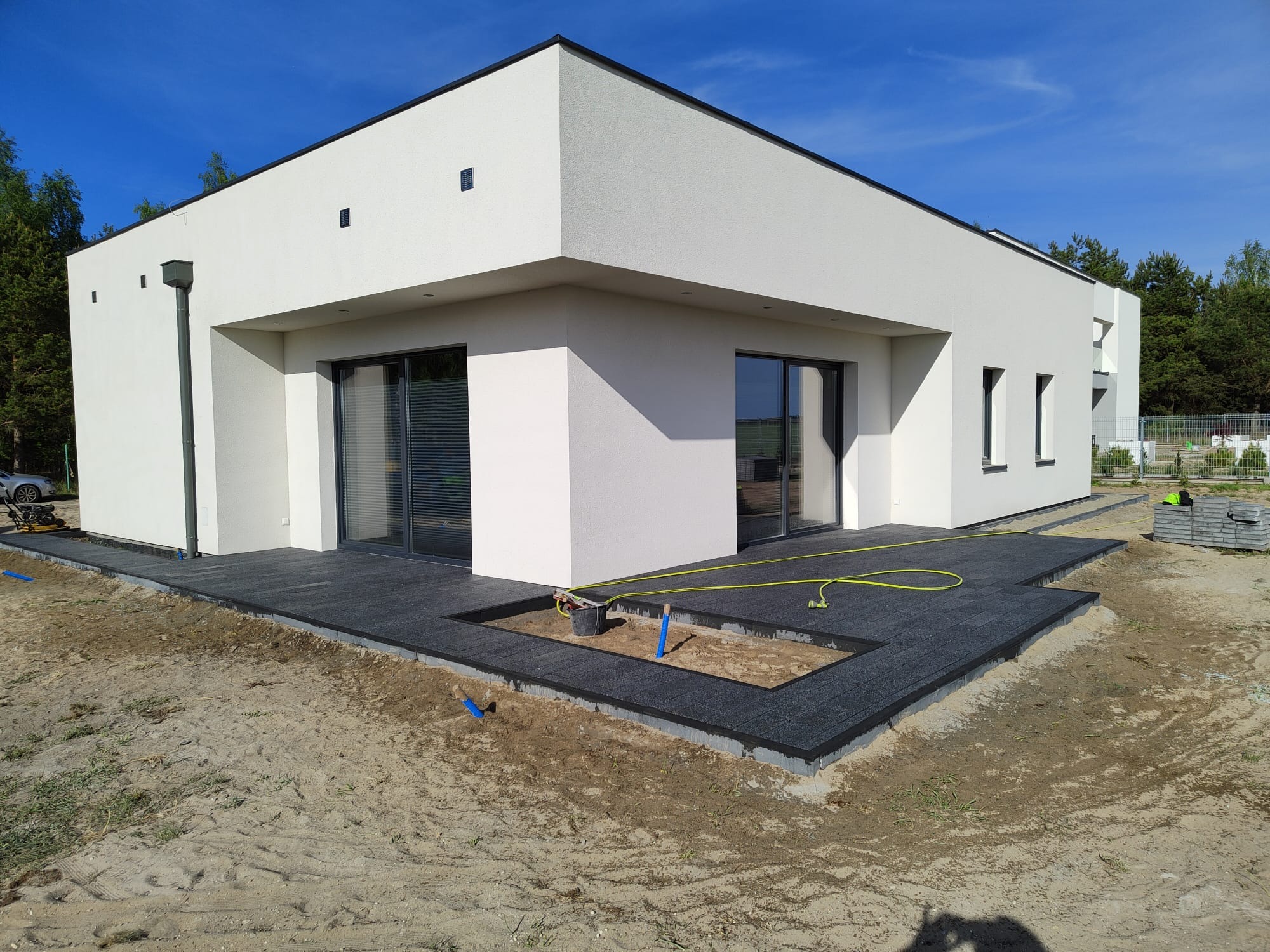 Nowoczesny dom z tarasem wyłożonym ciemnoszarą kostką, widoczne narzędzia i materiały budowlane, teren w trakcie prac ziemnych.