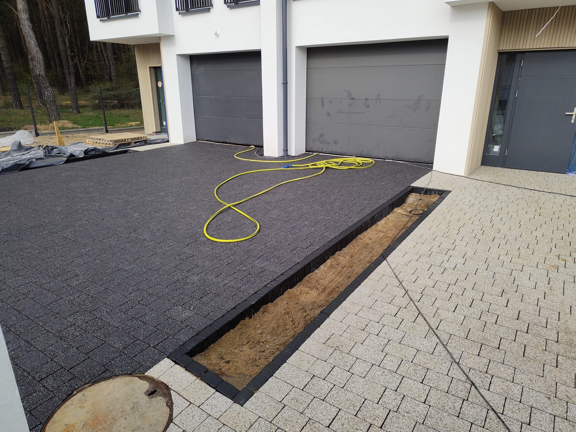 Układanie dwukolorowej kostki brukowej przed nowoczesnym budynkiem z dwoma garażami, widoczny fragment wykopu pod obrzeże, żółty wąż ogrodowy na ciemnej kostce.