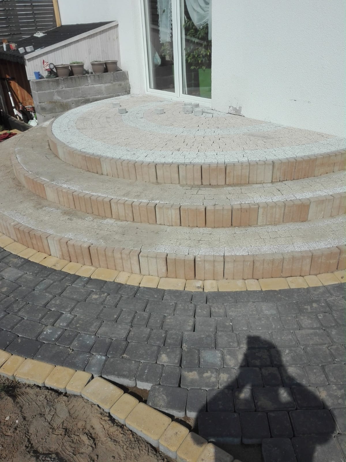 Półokrągłe schody wejściowe z kostki brukowej w trakcie układania, z widocznym fragmentem chodnika z ciemnej kostki obrzeżonego jaśniejszą. Na schodach widać jeszcze niezakończone fragmenty, a...