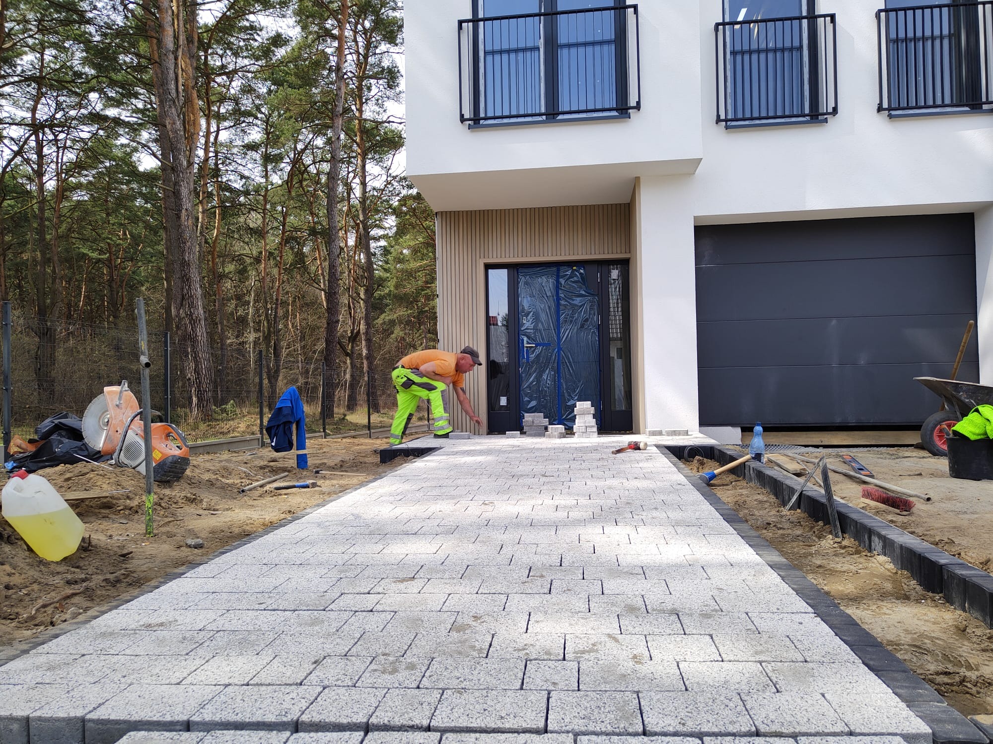 Pracownik w neonowej odzieży roboczej układa jasnoszarą kostkę brukową na podjeździe przed nowoczesnym domem z czarnymi oknami i bramą garażową. W tle las i narzędzia budowlane.