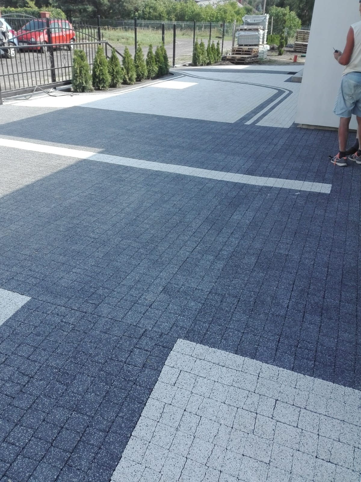 Ułożona, dwukolorowa nawierzchnia z kostki brukowej na podjeździe, z widocznymi białymi liniami i fragmentem budynku w tle.