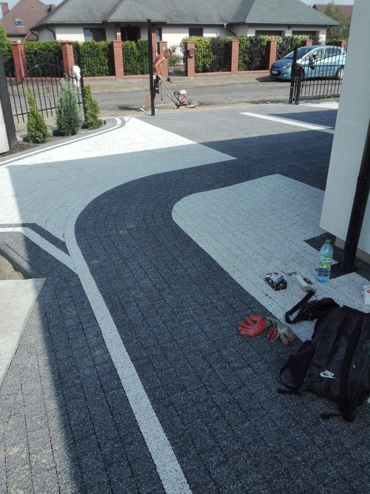 Ułożona kostka brukowa w odcieniach szarości z białymi akcentami, tworząca wzór na podjeździe. W tle mężczyzna używa maszyny do czyszczenia. Na pierwszym planie leżą robocze rękawice, plecak...