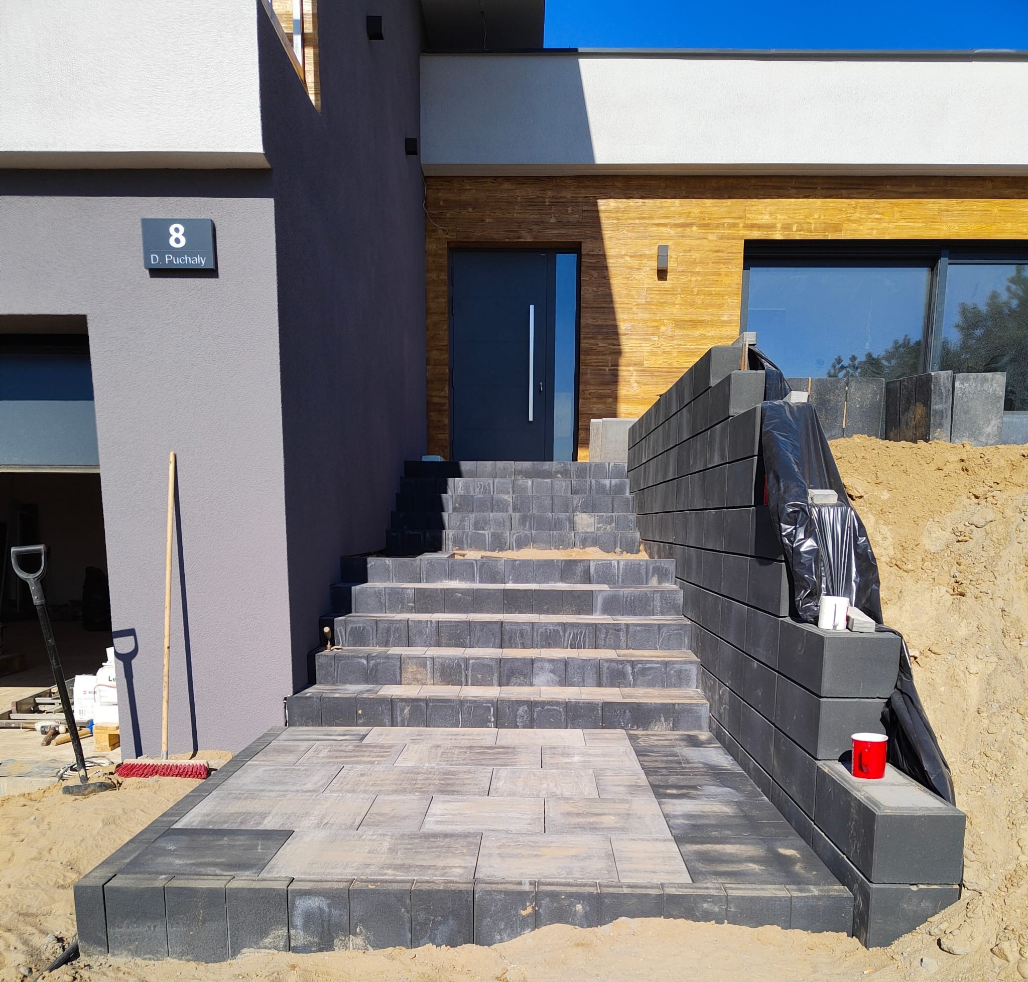 Schody wejściowe do domu wyłożone szarą kostką brukową, obok mur oporowy z bloczków, na którym stoi czerwony kubek.