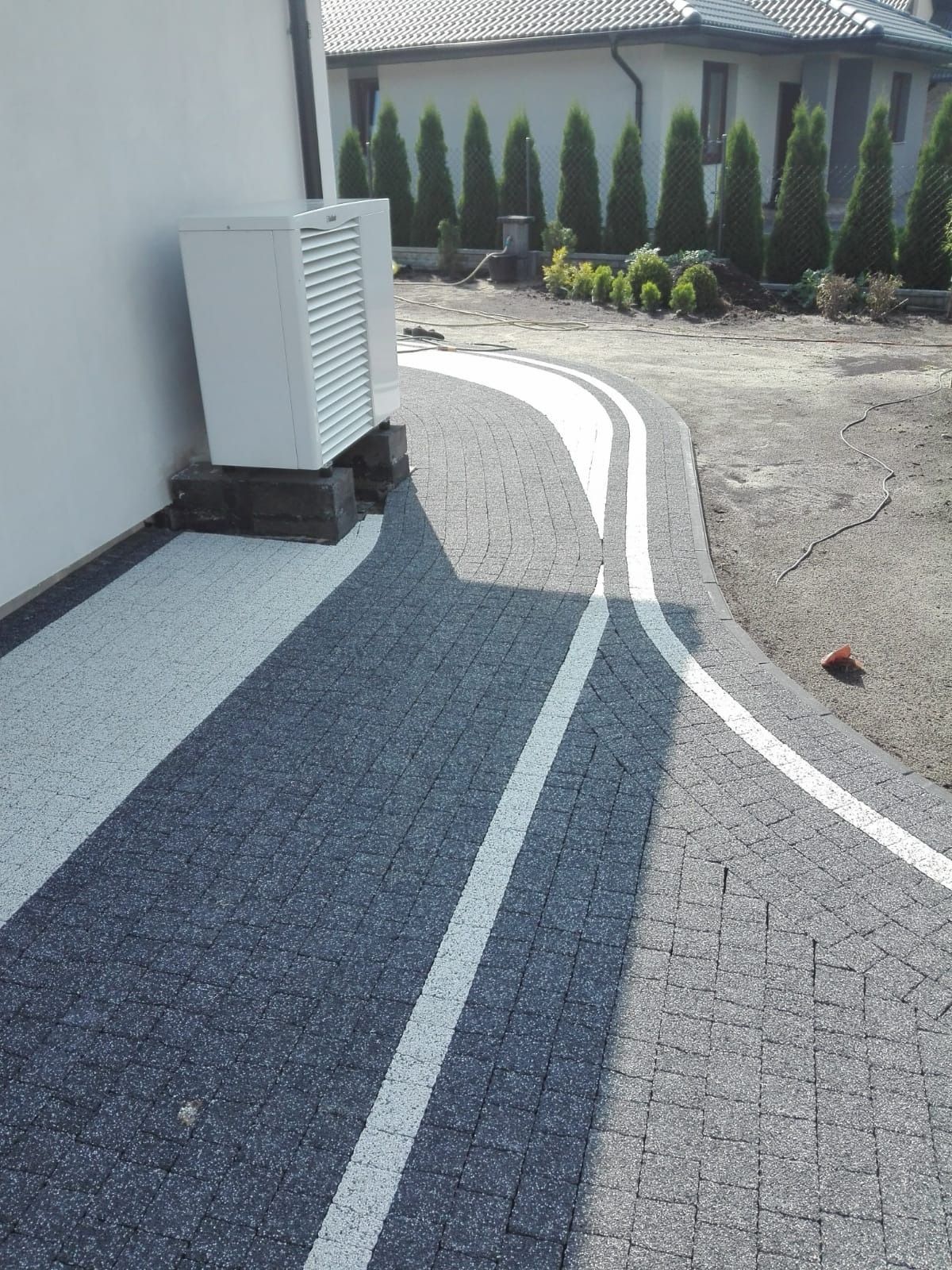 Ścieżka z szarej kostki brukowej z białymi pasami, prowadząca wzdłuż ściany budynku z zamontowaną jednostką klimatyzacji, w tle rząd tuj.