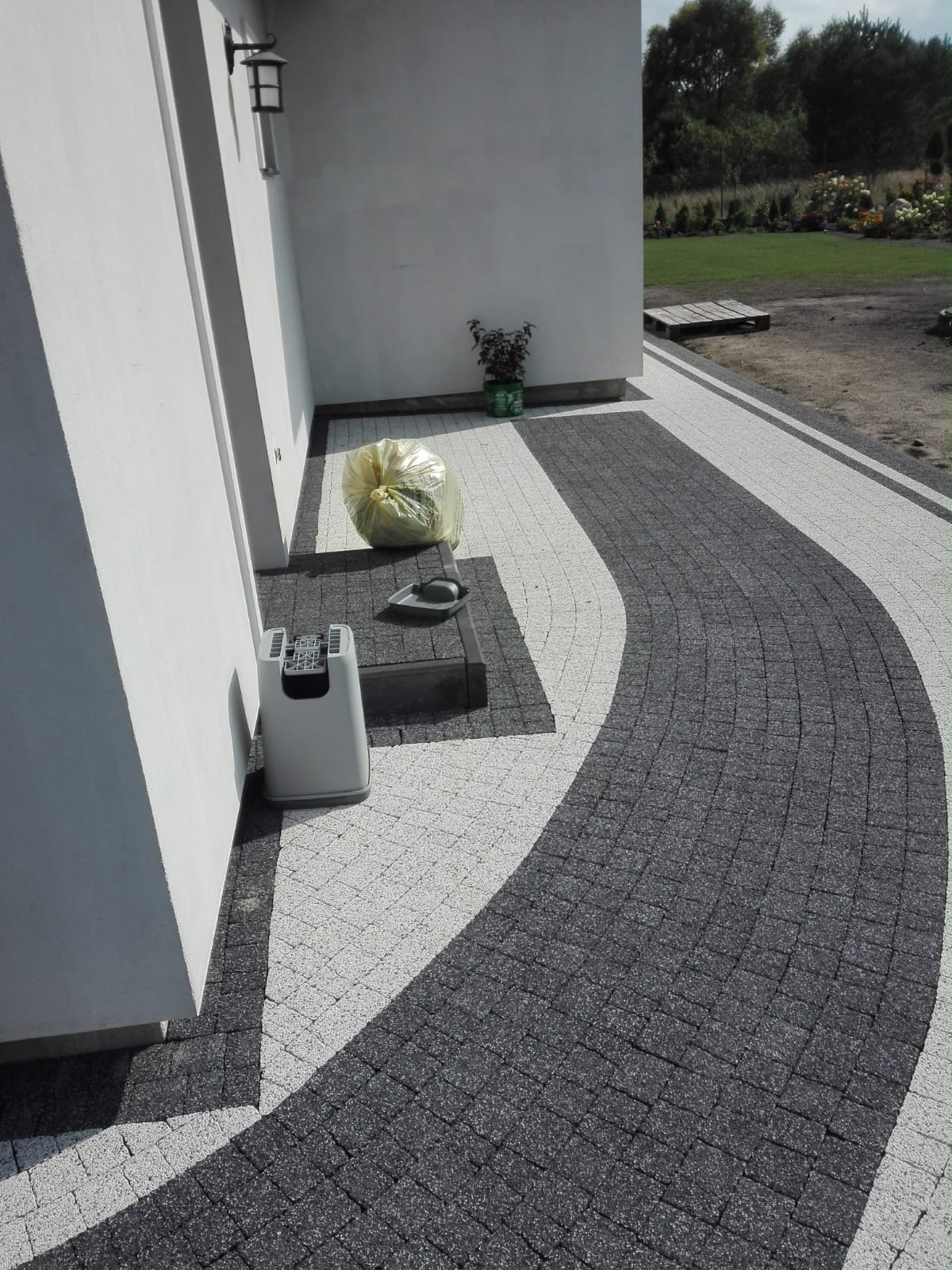 Nowo ułożona ścieżka z kostki brukowej w odcieniach szarości i bieli, zakręcająca łagodnym łukiem; widoczny fragment białej ściany budynku, worek z odpadami budowlanymi i narzędzia do sprzątania.