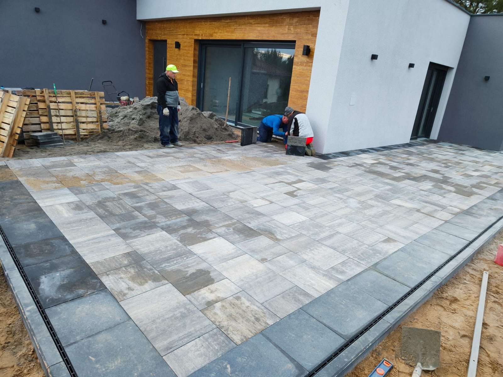 Układanie jasnoszarej kostki brukowej na posesji, widoczne palety z kostką, pracownicy w trakcie montażu obrzeży z odpływem liniowym.