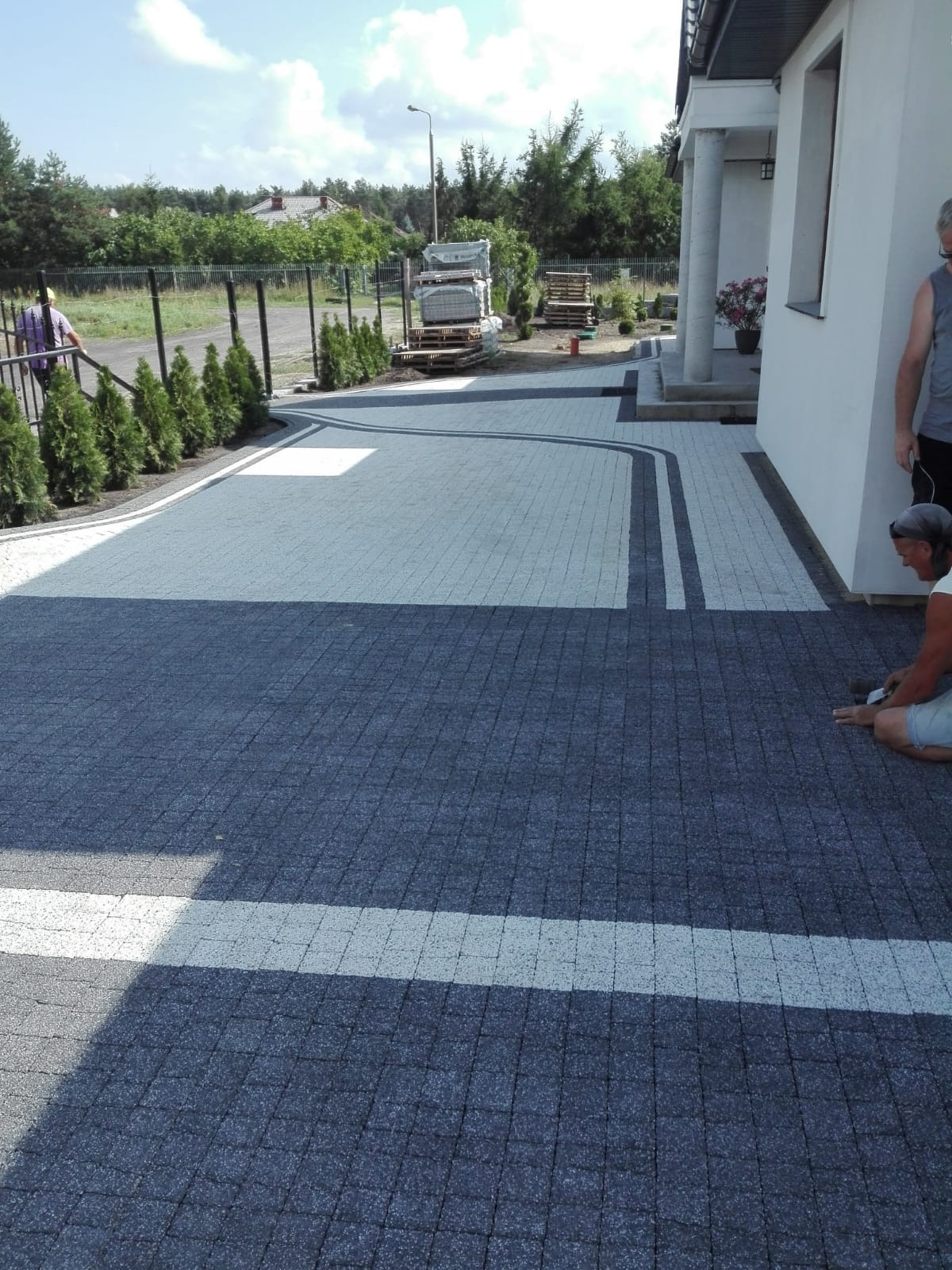 Ułożona dwukolorowa kostka brukowa z białymi pasami, pracownik na kolanach w trakcie układania, w tle zapasy kostki na paletach i dom z kolumnami.