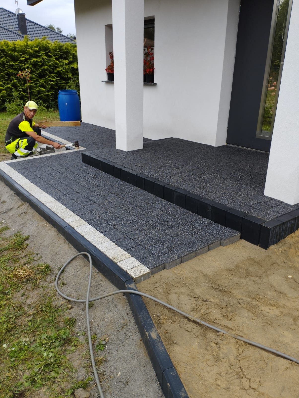 Pracownik w odblaskowej kamizelce układa ciemnoszarą kostkę brukową z jasnoszarą obwódką na podjeździe domu. Widoczny młotek brukarski, poziomica i częściowo ułożony fragment z czarnymi obrzeżami.