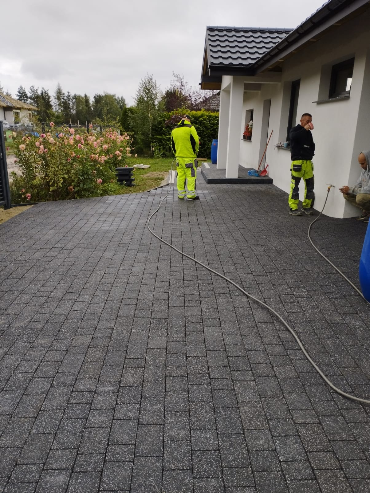 Ułożona szara kostka brukowa na podjeździe domu jednorodzinnego, widoczni trzej pracownicy w odblaskowych kombinezonach podczas prac wykończeniowych, jeden z nich czyści powierzchnię wodą...