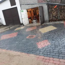 Ułożona kostka brukowa w odcieniach szarości, czerwieni i żółci, tworząca wzór na podjeździe przed garażem z otwartymi drzwiami, w tle widoczny dom i sterta drewna.