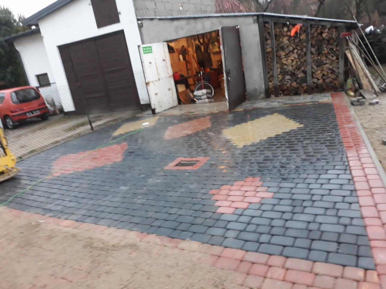 Ułożona kostka brukowa w odcieniach szarości, czerwieni i żółci, tworząca wzór na podjeździe przed garażem z otwartymi drzwiami, w tle widoczny dom i sterta drewna.