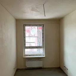 Pokój po nałożeniu gładzi cementowej; widoczne ślady po szpachlowaniu na suficie, okno z widokiem na rusztowanie, grzejnik zabezpieczony folią.