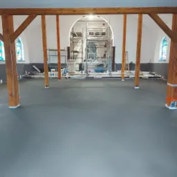 Wnętrze kościoła z świeżo wylaną, gładką, szarą posadzką; drewniane filary z pomarańczową taśmą zabezpieczającą, rusztowania w tle, okna witrażowe.