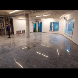 Usługi Remontowo-Budowlane Rafał Stawirej - Wnętrze biura po remoncie z szarą, marmurkową posadzką odbijającą światło, widoczne instalacje pod sufitem, kolumna w szarym oplocie oraz pracownik w niebieskiej kurtce przy instalacjach.