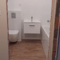 Wykończona łazienka z białymi płytkami ściennymi, podwieszaną toaletą, białą szafką z umywalką, wanną i podłogą imitującą drewno, widoczna zza progu z dekoracyjną, wzorzystą posadzką.