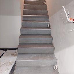 Usługi Remontowo-Budowlane Rafał Stawirej - Schody wyłożone szarymi płytkami z metalowymi listwami, ściany w trakcie malowania, widoczne kable elektryczne i wiadro z farbą na górze schodów.
