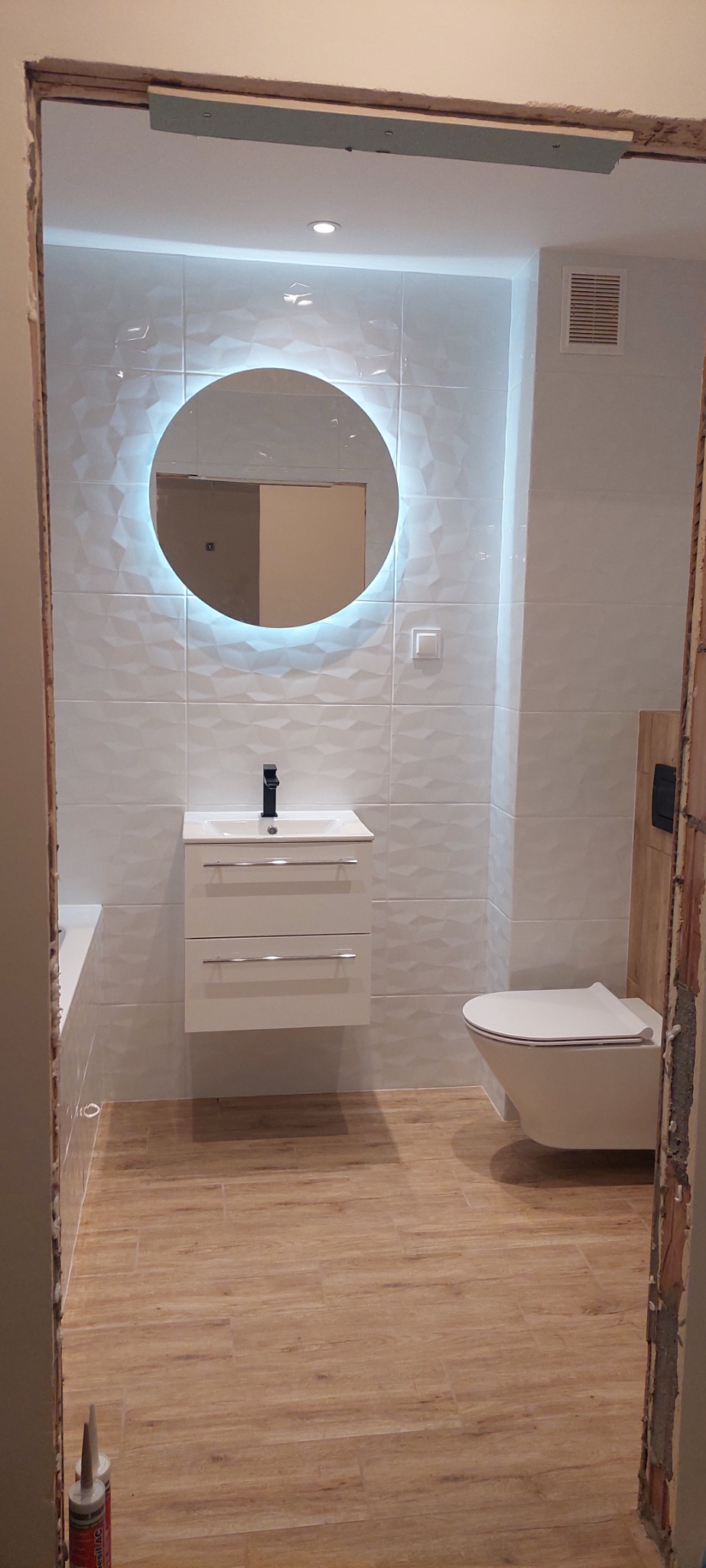 Nowoczesna łazienka z podświetlanym okrągłym lustrem, umywalką z szafką, wiszącą toaletą i drewnopodobną podłogą, widoczna przez surową framugę drzwi.