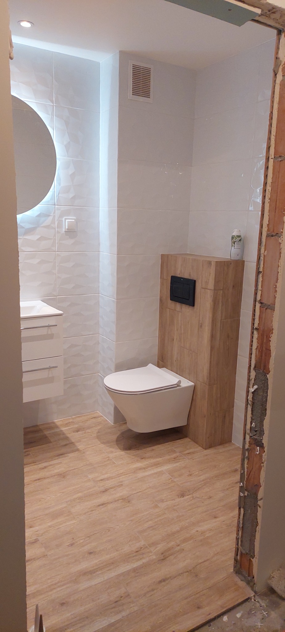 Nowoczesna łazienka z podwieszaną toaletą, umywalką z szafką i okrągłym lustrem z podświetleniem LED, wykończona białymi płytkami z trójwymiarowym wzorem i panelami podłogowymi imitującymi drewno...