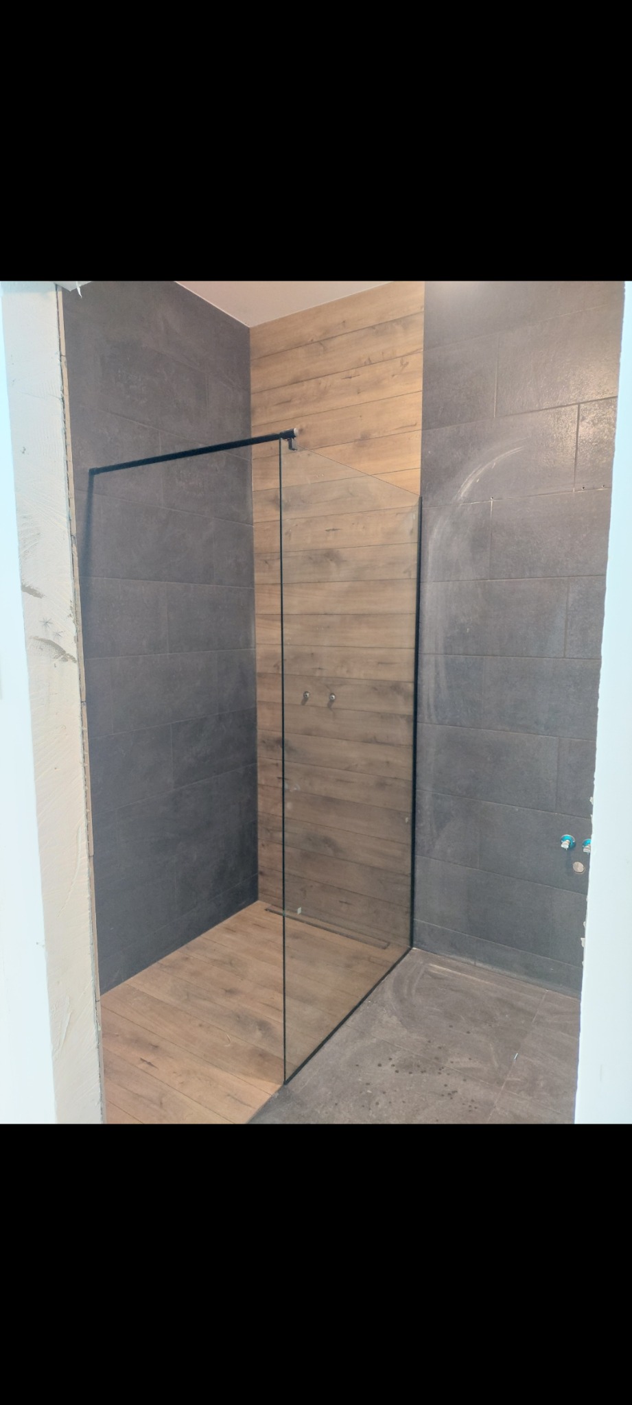 Nowoczesna kabina prysznicowa typu walk-in z czarnymi profilami, drewnopodobne płytki na ścianie i podłodze wewnątrz kabiny, szare płytki na pozostałej części ścian, widoczne niedokończone prace...