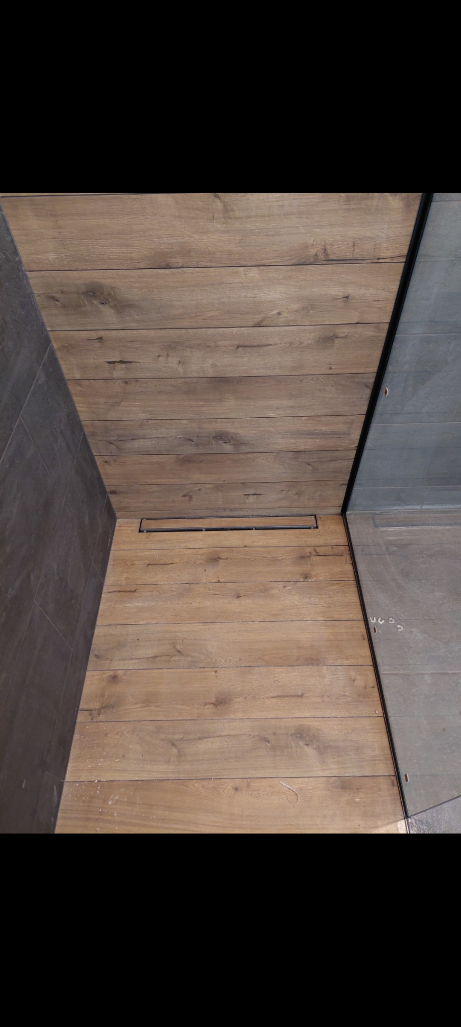 Wykończona podłoga w kabinie prysznicowej z drewnopodobnych płytek, z liniowym odpływem, widok z góry, szara ściana i szklana tafla w tle.