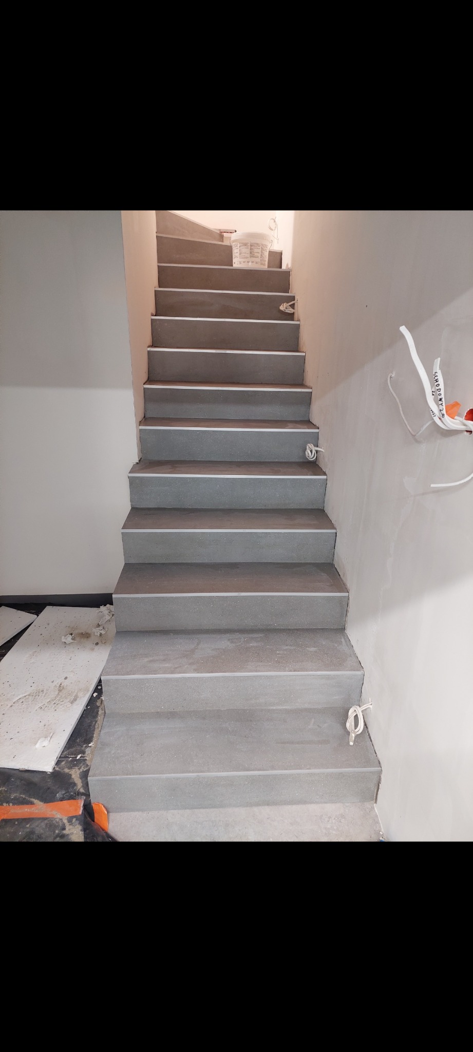 Schody wyłożone szarą płytką imitującą beton, z widocznym fragmentem instalacji elektrycznej na ścianie i wiadrem po farbie na górze schodów.