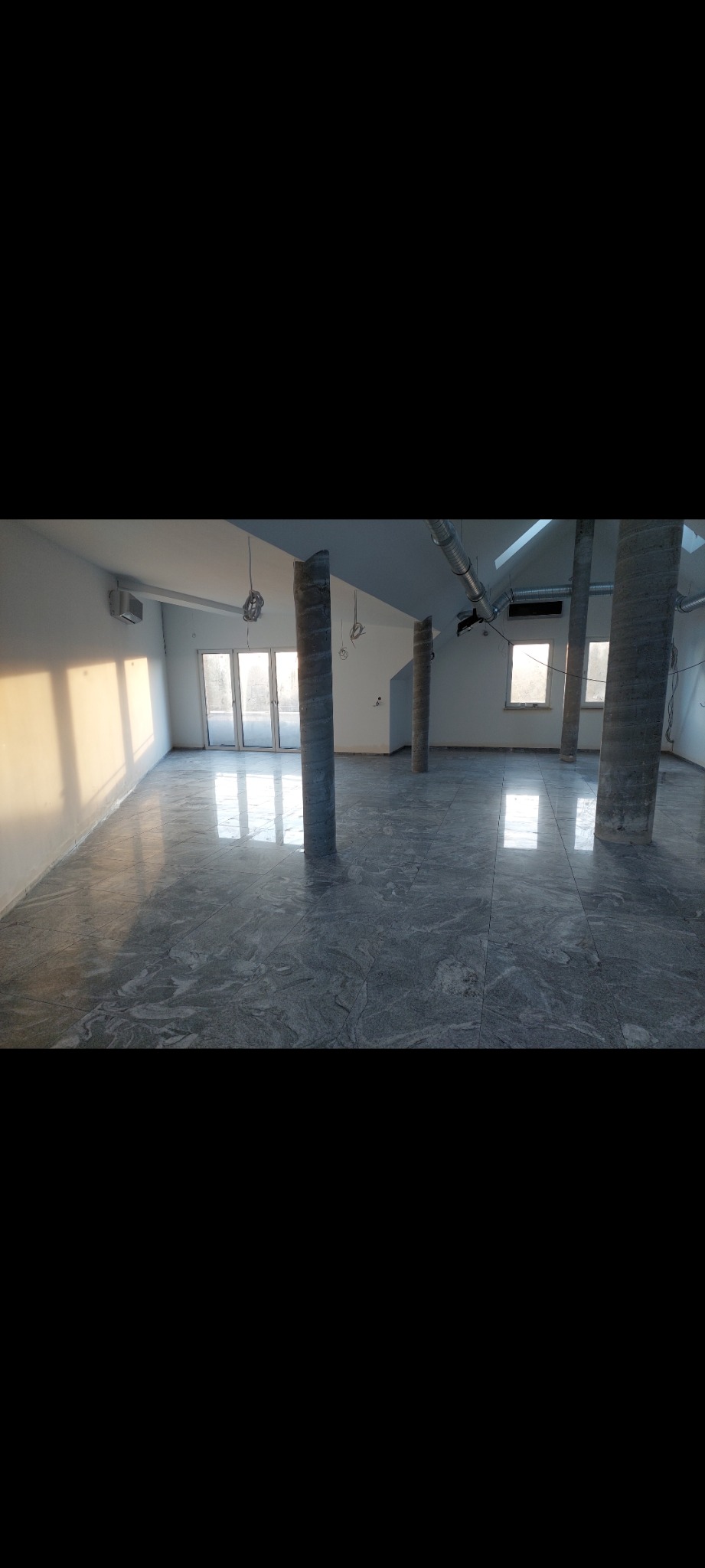 Wnętrze biurowe w trakcie wykańczania: szara marmurkowa posadzka, betonowe kolumny, widoczne instalacje pod sufitem, wpadające światło słoneczne przez okna.