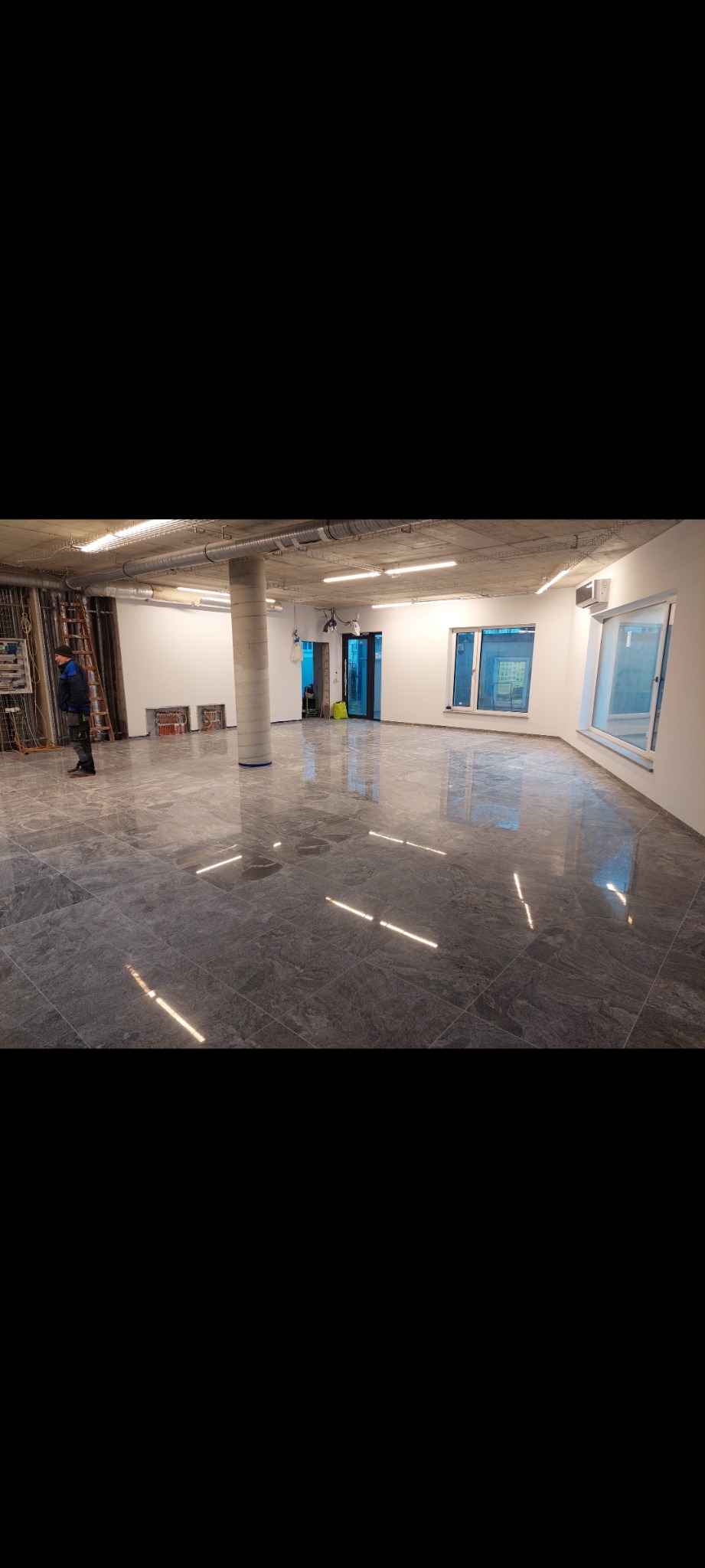 Wnętrze biura po remoncie z szarą, marmurkową posadzką odbijającą światło, widoczne instalacje pod sufitem, kolumna w szarym oplocie oraz pracownik w niebieskiej kurtce przy instalacjach.