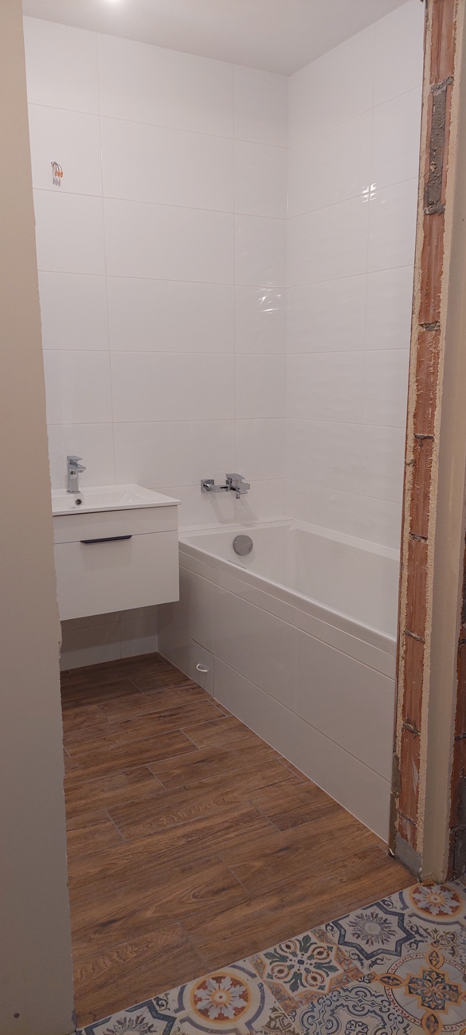 Nowoczesna łazienka z białą armaturą, drewnopodobną podłogą i fragmentem ściany z odsłoniętą cegłą w trakcie wykończenia.