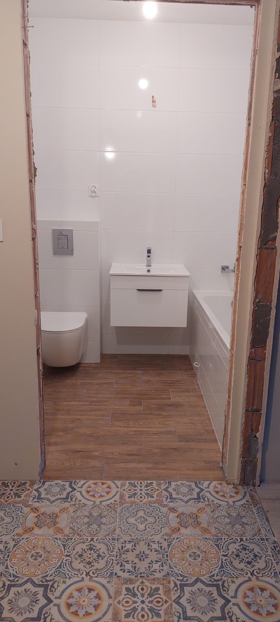 Wykończona łazienka z białymi płytkami ściennymi, podwieszaną toaletą, białą szafką z umywalką, wanną i podłogą imitującą drewno, widoczna zza progu z dekoracyjną, wzorzystą posadzką.