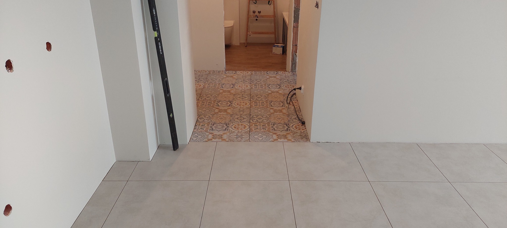 Fragment wnętrza po remoncie: ułożona jasna, kwadratowa podłoga z płytek ceramicznych przechodzi w dekoracyjną posadzkę w przejściu do łazienki, widoczna poziomica oparta o ścianę, ściany białe...