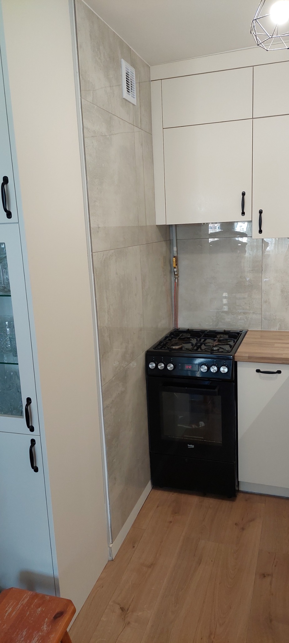 Wykończona kuchnia z beżowymi płytkami imitującymi beton, zabudową meblową w odcieniach bieli, czarną kuchenką gazową i drewnianą podłogą.