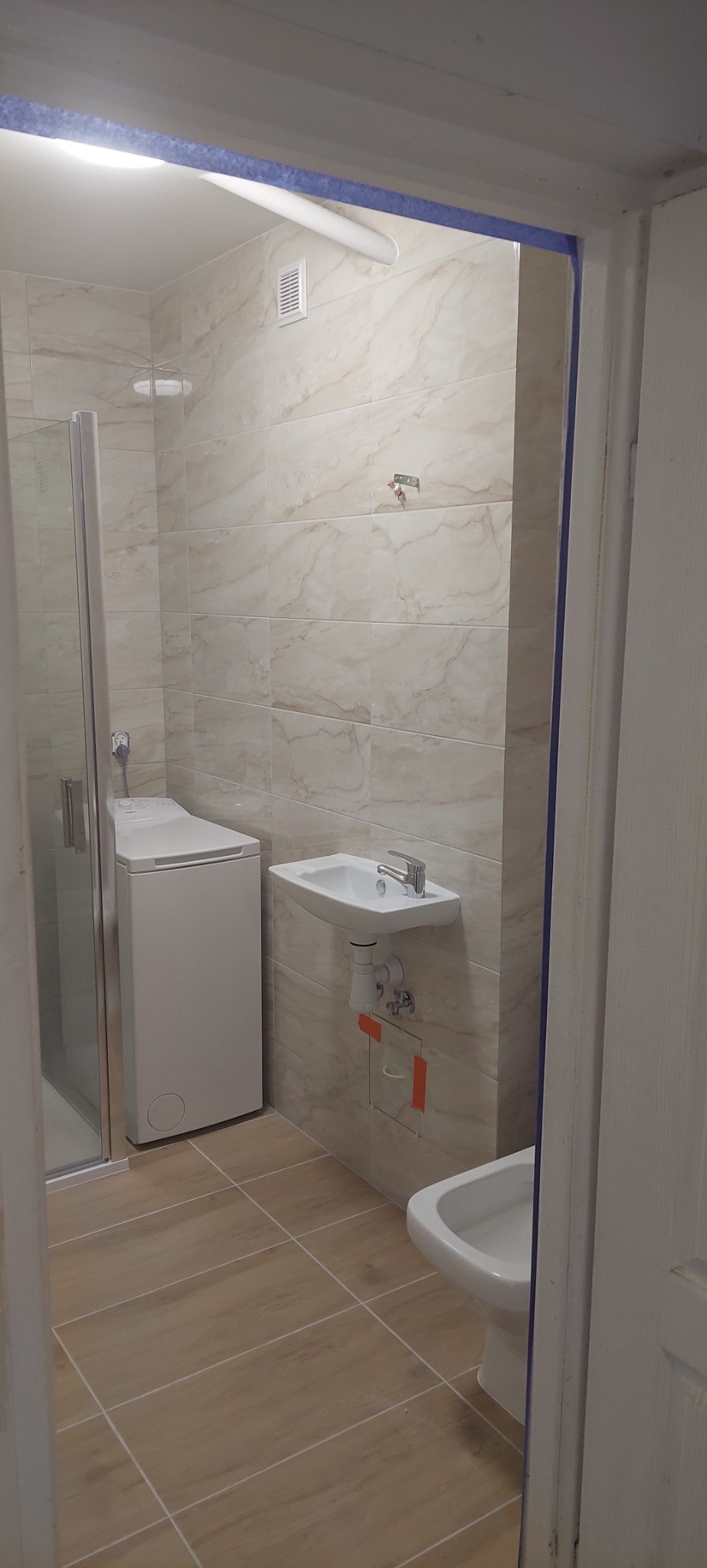 Wykończona łazienka z beżowymi płytkami imitującymi marmur, pralką pod prysznicem, umywalką na ścianie i bidetem, widok zza futryny.