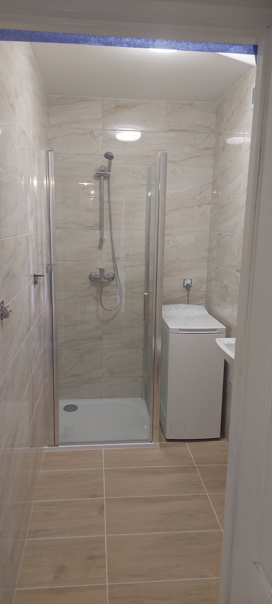 Wykończona łazienka z kabiną prysznicową z przezroczystego szkła, beżowymi płytkami imitującymi marmur na ścianach, brązowymi płytkami podłogowymi, białą pralką ładowaną od góry i fragmentem umywalki.