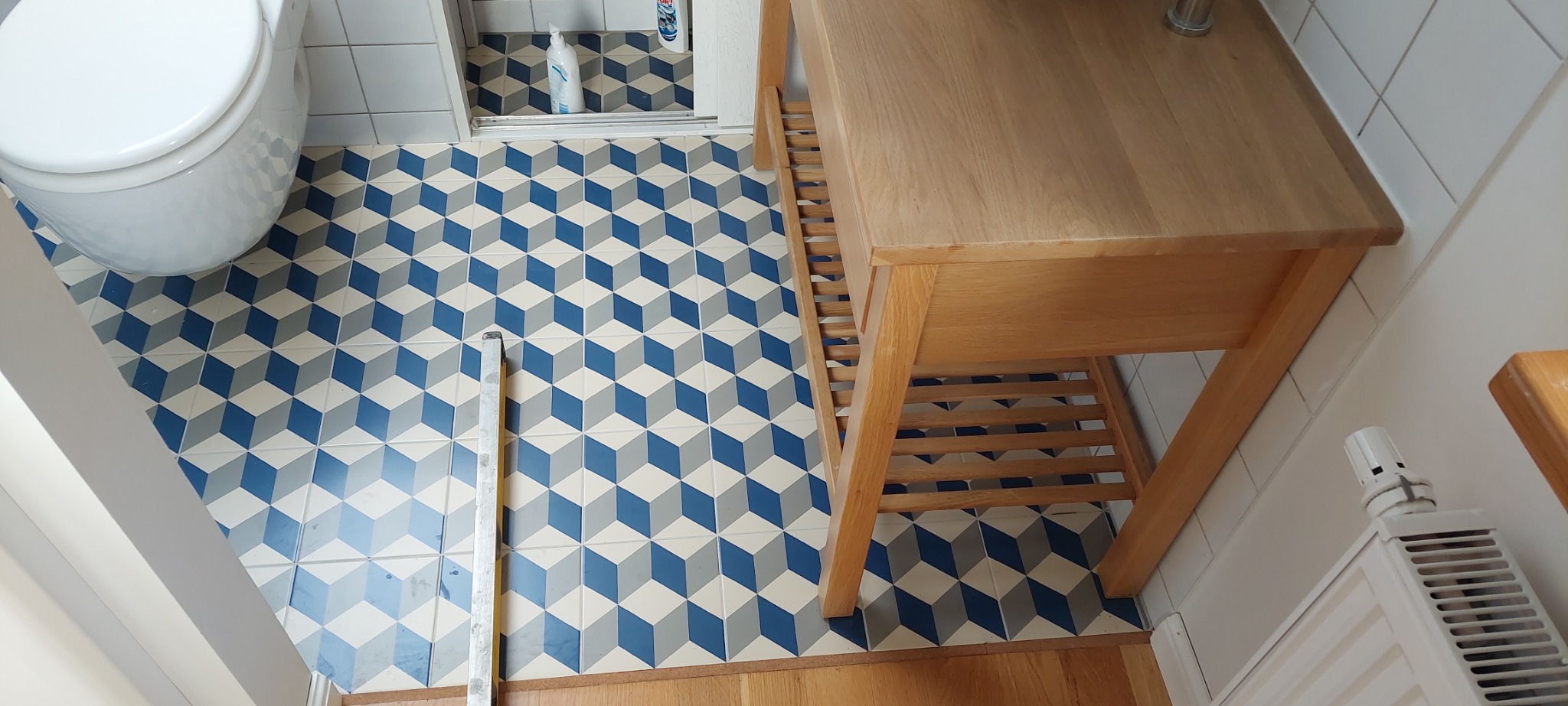 Wzorzyste, niebiesko-szare płytki podłogowe w łazience z efektem 3D, widoczny fragment toalety, drewniana szafka pod umywalkę i biały grzejnik.