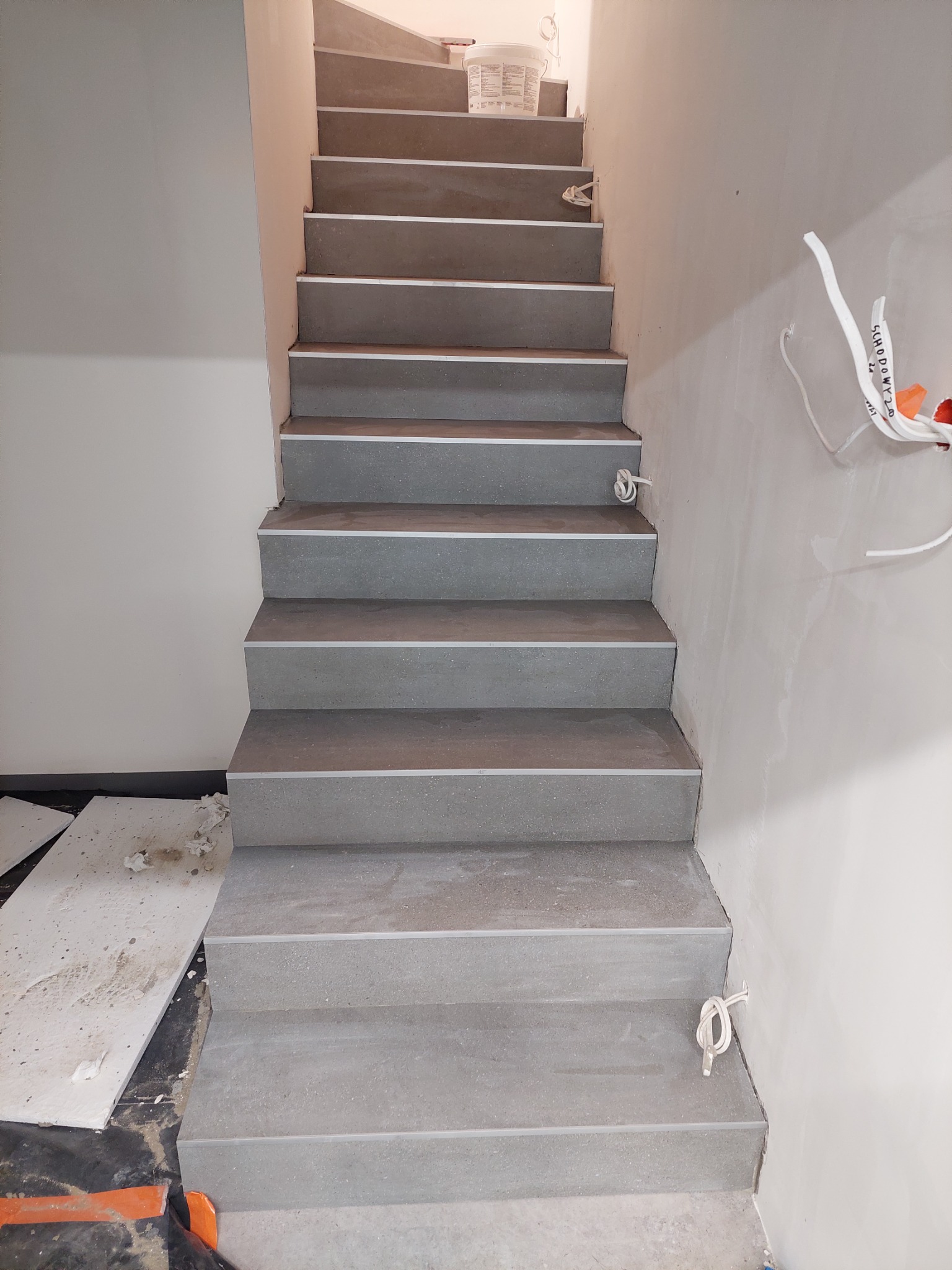 Schody wyłożone szarymi płytkami z metalowymi listwami, ściany w trakcie malowania, widoczne kable elektryczne i wiadro z farbą na górze schodów.