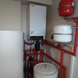 Kompletna instalacja grzewcza Viessmann z czerwonymi rurami, zasobnikiem, kotłem i naczyniami wzbiorczymi w kotłowni o szarych ścianach i żółtym przejściu do innego pomieszczenia.