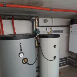 Dwa zbiorniki ciepłej wody użytkowej, Atlantic i inny, w kotłowni, widoczne rury w izolacji termicznej, manometry, zawory i instalacja elektryczna.