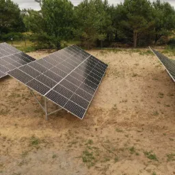 Instalacja fotowoltaiczna na gruncie z panelami ustawionymi pod kątem, widoczne metalowe konstrukcje wsporcze, piaszczyste podłoże z rzadką roślinnością, w tle drzewa i zabudowania.