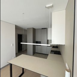 Inside Home Building - Nowoczesna, jasna kuchnia w odcieniach szarości i beżu z minimalistycznym oświetleniem LED. Widoczne szafki, blat i stół z czarnymi nogami. W tle otwarte drzwi.