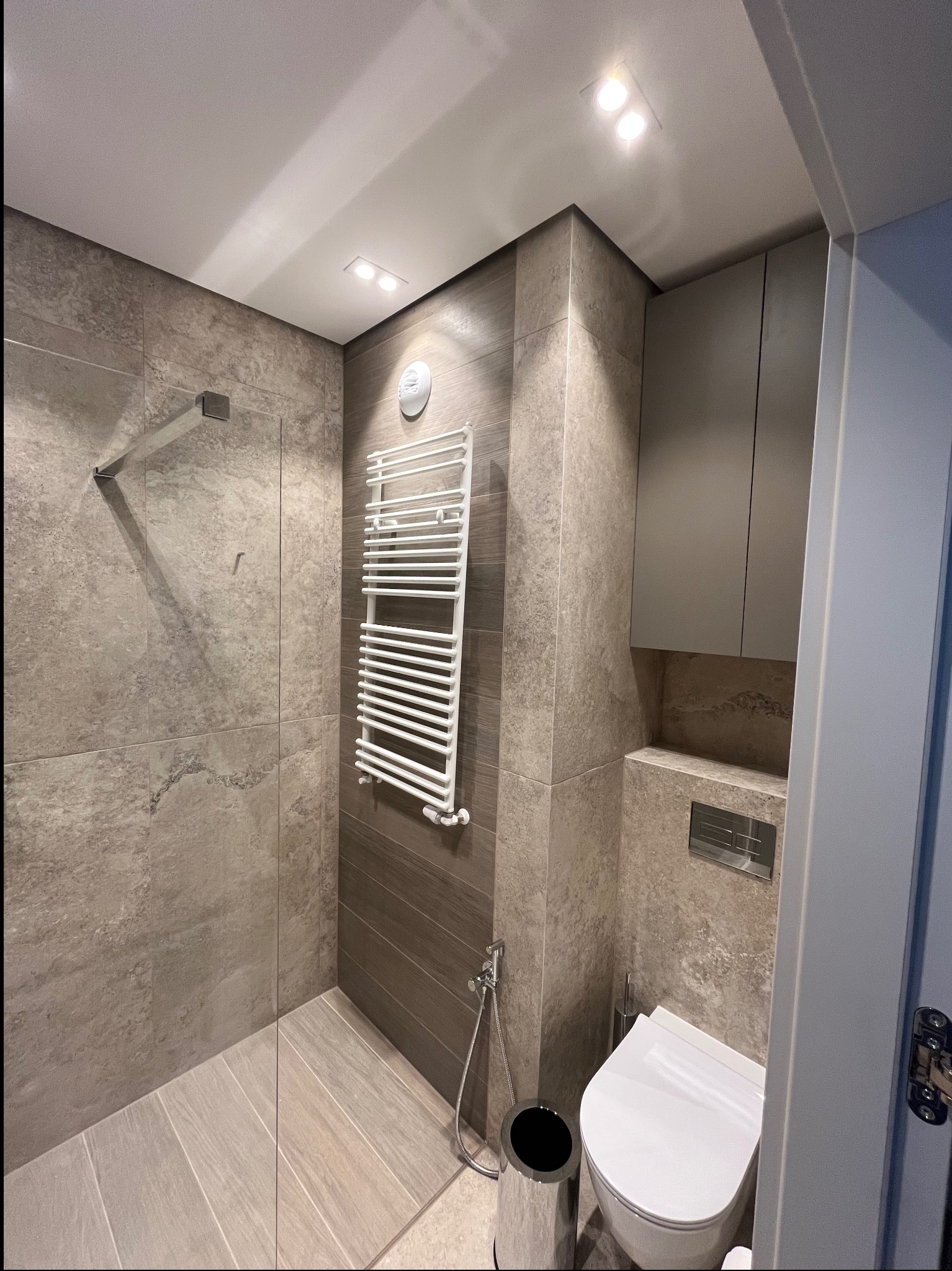 Nowoczesna łazienka z kabiną prysznicową typu walk-in, grzejnikiem drabinkowym i podwieszaną toaletą. Wykończenie w odcieniach szarości, płytki imitujące kamień. Oświetlenie punktowe.